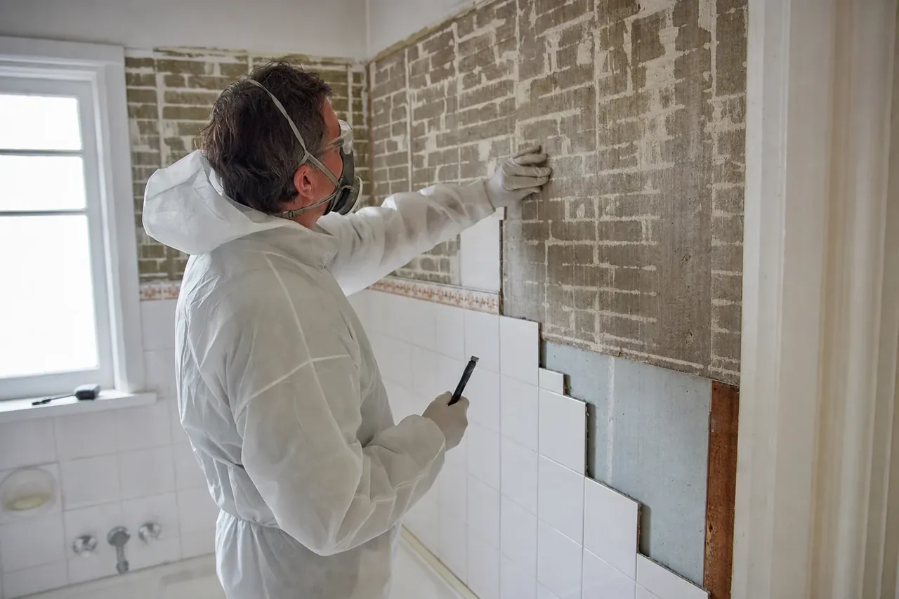 Licensed asbestos assessor inspecting old wall sheeting behind partially removed bathroom tiles in an older Perth home